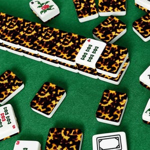 May include: A collection of Mahjong tiles with a tortoiseshell pattern on a green surface. The tiles are white with various symbols and numbers in red and green. Some tiles feature floral designs.