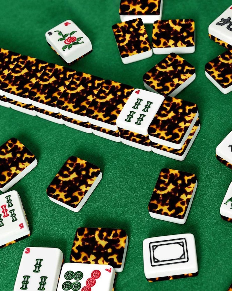 May include: A collection of Mahjong tiles with a tortoiseshell pattern on a green surface. The tiles are white with various symbols and numbers in red and green. Some tiles feature floral designs.