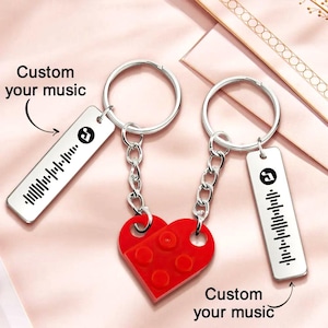 Puede incluir: Dos llaveros plateados con etiquetas rectangulares que muestran una onda de sonido musical y el texto "Custom your music". Un dije rojo en forma de corazón hecho de bloques entrelazados conecta los llaveros. El fondo es rosa claro.