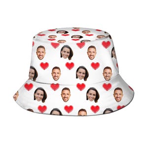 May include: White bucket hat with a repeating pattern of faces and red hearts. The faces are of a man and a woman. This casual hat is a stylish accessory, suitable for sun protection.