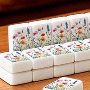 K&ouml;nnte beinhalten: Ein Set aus wei&szlig;en, rechteckigen Mahjong-Spielsteinen mit einem farbenfrohen Aufdruck verschiedener Wildblumen, darunter rosa, rote, gelbe und blaue Bl&uuml;ten mit gr&uuml;nen St&auml;ngeln und Bl&auml;ttern auf der Vorderseite jedes einzelnen Spielsteins.