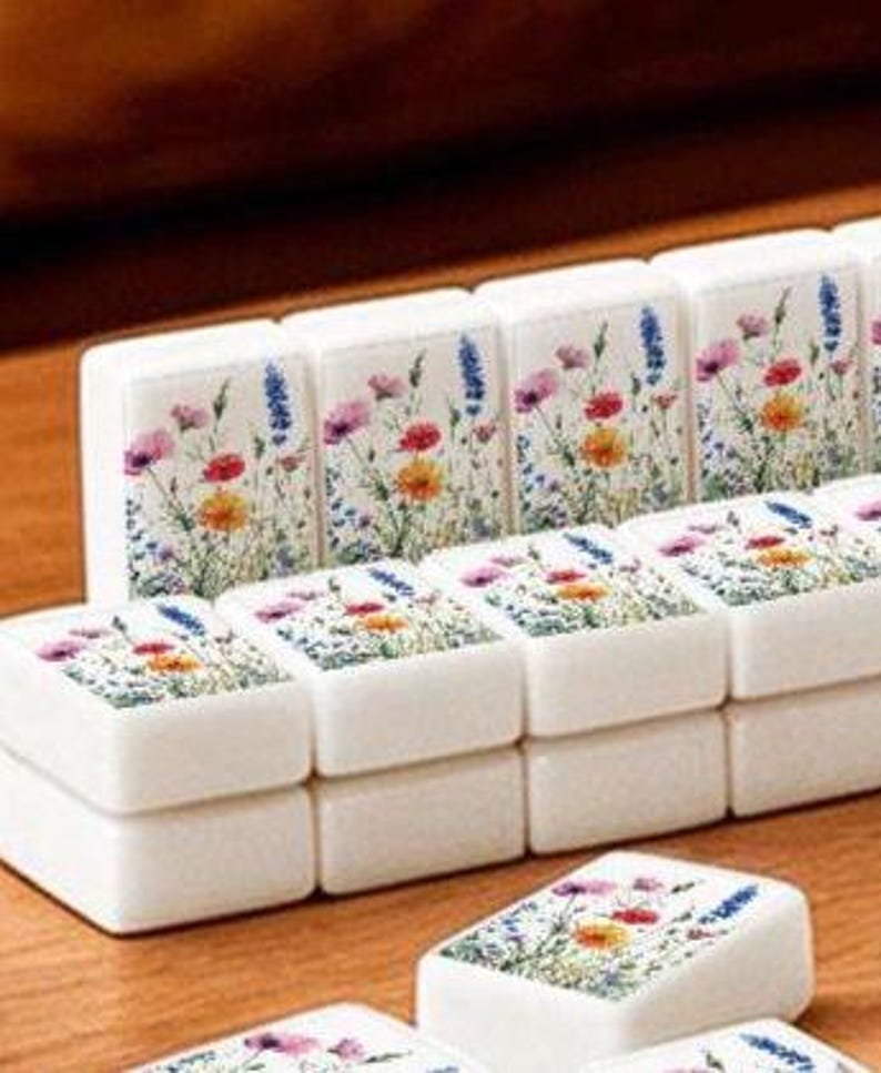K&ouml;nnte beinhalten: Ein Set aus wei&szlig;en, rechteckigen Mahjong-Spielsteinen mit einem farbenfrohen Aufdruck verschiedener Wildblumen, darunter rosa, rote, gelbe und blaue Bl&uuml;ten mit gr&uuml;nen St&auml;ngeln und Bl&auml;ttern auf der Vorderseite jedes einzelnen Spielsteins.
