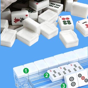 May include: A white Mahjong set with various symbols and numbers, including red and green markings. The tiles are displayed in a clear plastic rack and a beige cloth bag. The game set is on a blue background.