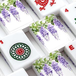 Pode incluir: Pe&ccedil;as de Mahjong brancas com v&aacute;rios desenhos. Algumas pe&ccedil;as apresentam flores de glic&iacute;nia roxas com folhas verdes, enquanto outras exibem um drag&atilde;o vermelho ou padr&otilde;es circulares verdes e vermelhos. As pe&ccedil;as est&atilde;o dispostas em uma caixa.