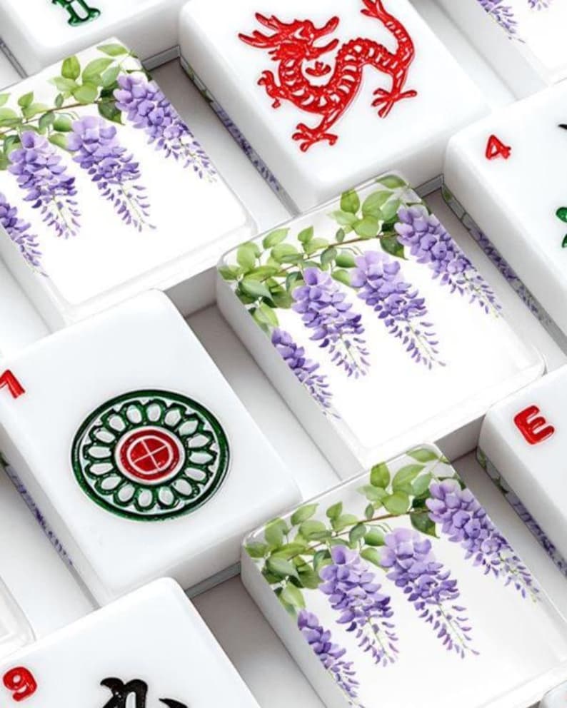 Pode incluir: Pe&ccedil;as de Mahjong brancas com v&aacute;rios desenhos. Algumas pe&ccedil;as apresentam flores de glic&iacute;nia roxas com folhas verdes, enquanto outras exibem um drag&atilde;o vermelho ou padr&otilde;es circulares verdes e vermelhos. As pe&ccedil;as est&atilde;o dispostas em uma caixa.