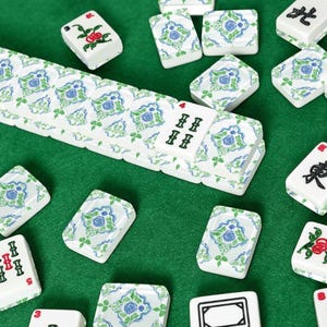 Puede incluir: Azulejos de Mahjong blancos con dise&ntilde;os florales azules y verdes dispersos sobre una superficie verde. Algunas fichas muestran caracteres chinos y n&uacute;meros. Las fichas est&aacute;n dispuestas, sugiriendo un juego en curso.