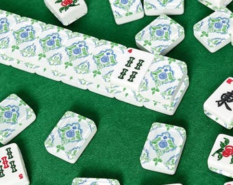 May include: A collection of white Mahjong tiles with blue and green floral patterns, scattered on a green surface. Some tiles display Chinese characters and numbers. The tiles are rectangular and have a glossy finish, suggesting a classic board game.