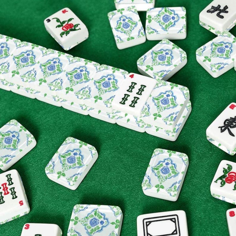 Puede incluir: Azulejos de Mahjong blancos con dise&ntilde;os florales azules y verdes dispersos sobre una superficie verde. Algunas fichas muestran caracteres chinos y n&uacute;meros. Las fichas est&aacute;n dispuestas, sugiriendo un juego en curso.