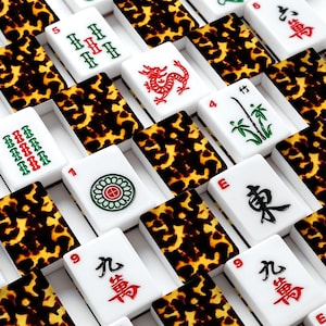 May include: A close-up of a Mahjong set with white tiles featuring red, green, and black symbols and numbers. The tiles are arranged in a grid pattern, interspersed with brown and yellow patterned tiles.