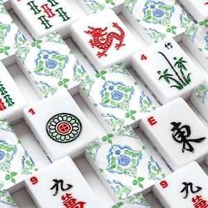 Puede incluir: Primer plano de un juego de Mahjong con fichas blancas con s&iacute;mbolos y n&uacute;meros rojos, verdes y negros. Algunas fichas muestran un drag&oacute;n rojo, bamb&uacute; y dise&ntilde;os florales. Las fichas est&aacute;n dispuestas en una caja con un patr&oacute;n floral.