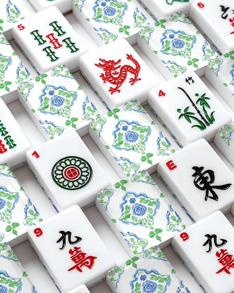 Puede incluir: Primer plano de un juego de Mahjong con fichas blancas con s&iacute;mbolos y n&uacute;meros rojos, verdes y negros. Algunas fichas muestran un drag&oacute;n rojo, bamb&uacute; y dise&ntilde;os florales. Las fichas est&aacute;n dispuestas en una caja con un patr&oacute;n floral.