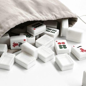 May include: A collection of white Mahjong tiles with red and green markings spilling from a beige drawstring bag. The tiles are rectangular with various symbols and characters.