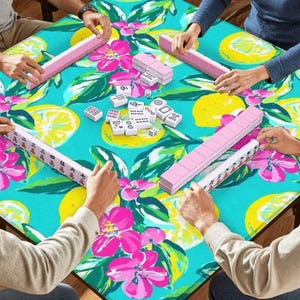 Puede incluir: Un grupo de personas jugando al mahjong en una mesa cuadrada. La mesa tiene un estampado floral turquesa con limones y flores rosas. Las fichas de mahjong son blancas con marcas negras y los atriles son rosas.