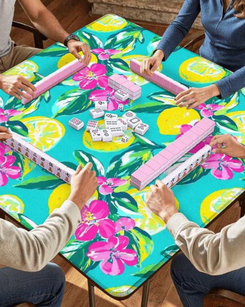 Puede incluir: Un grupo de personas jugando al mahjong en una mesa cuadrada. La mesa tiene un estampado floral turquesa con limones y flores rosas. Las fichas de mahjong son blancas con marcas negras y los atriles son rosas.