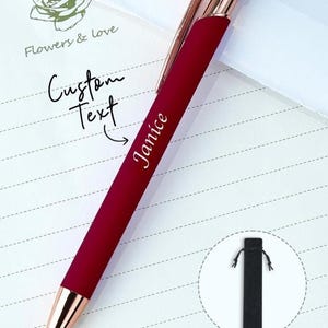 Op de afbeelding: Een bordeauxrode pen met goudkleurige accenten en de naam "Janice" aan de zijkant. De pen wordt getoond op gelijnd papier met de woorden "Flowers & Love" en "Custom Text". Een zwarte pennenetui is ook afgebeeld.