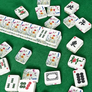 May include: A collection of white Mahjong tiles with various symbols and floral designs, scattered on a green surface. Some tiles feature red, pink, and yellow flowers. Other tiles have Chinese characters and pips.