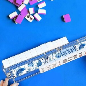 May include: A complete Mahjong set with white and purple tiles, some scattered on a blue surface. The tile case features a blue and white floral design with koi fish. The word "ARDE" is visible on the case.