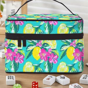 May include: A rectangular cosmetic bag with a black handle and zip. The bag displays a bright pattern of yellow lemons, pink flowers, and green leaves on a turquoise background. Mahjong tiles and dice are in front of the bag.
