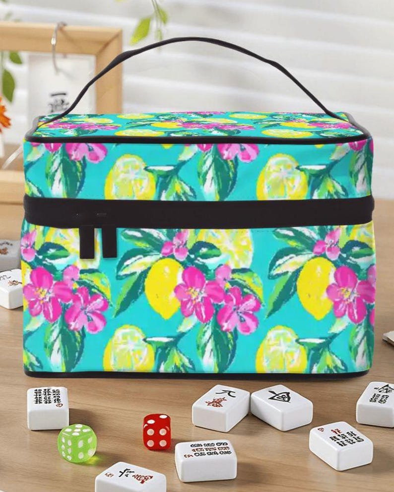 May include: A rectangular cosmetic bag with a black handle and zip. The bag displays a bright pattern of yellow lemons, pink flowers, and green leaves on a turquoise background. Mahjong tiles and dice are in front of the bag.