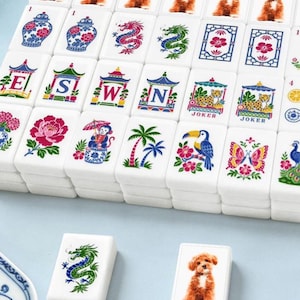 May include: A set of white rectangular mahjong tiles featuring colorful illustrations of brown puppies, blue and white vases, dragons, flowers, palm trees, toucans, butterflies, and tiles marked with the letters E, S, W, N and the word JOKER.