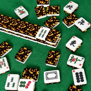 Op de afbeelding: Een verzameling ivoor- en schildpadpatroon Mahjong-tegels verspreid op een groene ondergrond. De tegels tonen verschillende symbolen, cijfers en bloemmotieven. Sommige tegels zijn gestapeld, andere verspreid, wat de spelcomponenten laat zien.