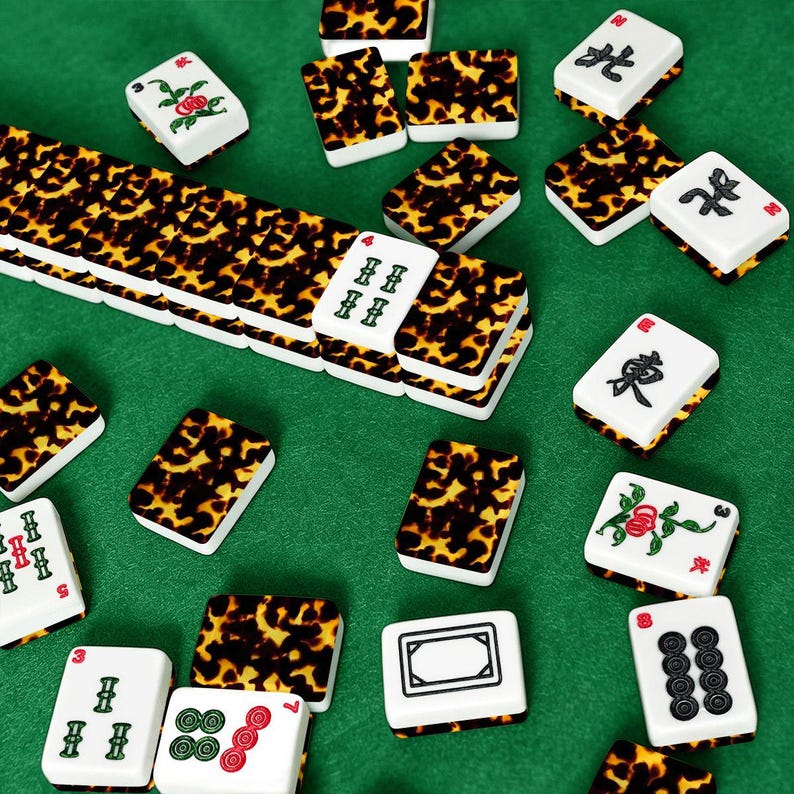 Op de afbeelding: Een verzameling ivoor- en schildpadpatroon Mahjong-tegels verspreid op een groene ondergrond. De tegels tonen verschillende symbolen, cijfers en bloemmotieven. Sommige tegels zijn gestapeld, andere verspreid, wat de spelcomponenten laat zien.
