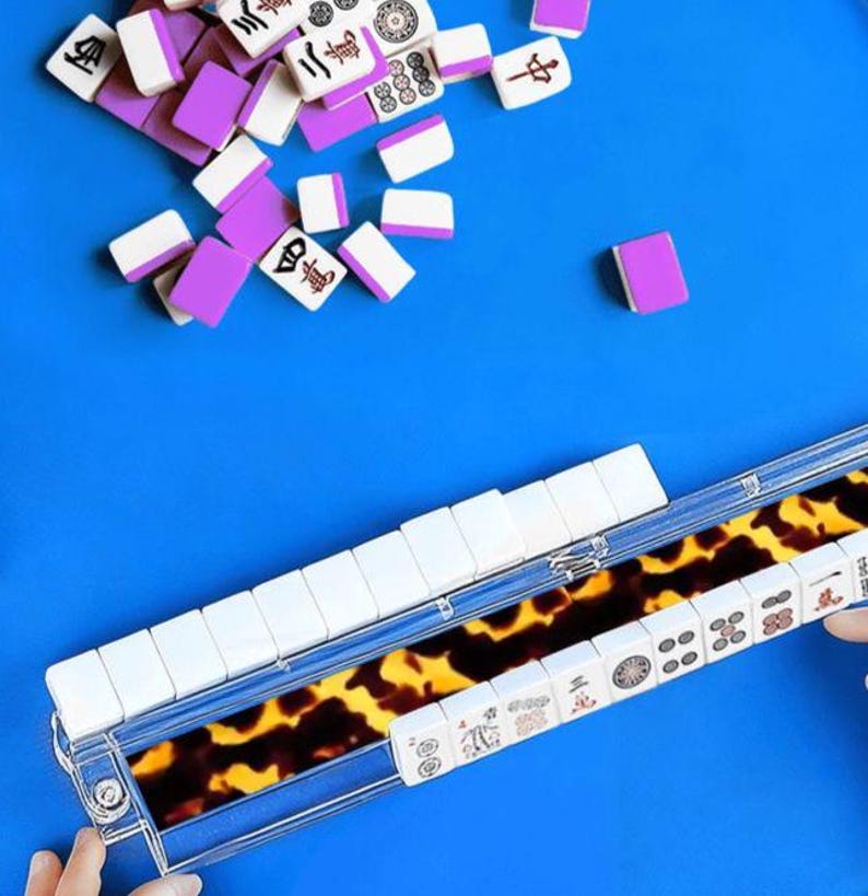 May include: A complete Mahjong set with white tiles and purple accents, scattered on a bright blue surface. The tiles are arranged in a clear case with a tortoiseshell pattern. The game set includes a variety of symbols and characters.