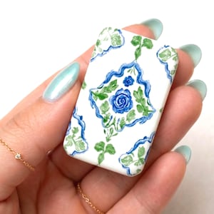 May include: A rectangular, white ceramic tile with a blue floral design. The design features a central blue rose surrounded by green leaves and a blue border. The tile is held in a hand with light blue nail polish.