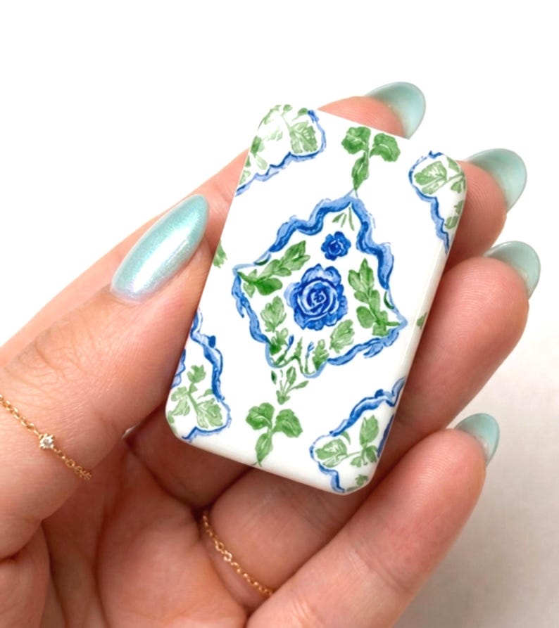 May include: A rectangular, white ceramic tile with a blue floral design. The design features a central blue rose surrounded by green leaves and a blue border. The tile is held in a hand with light blue nail polish.