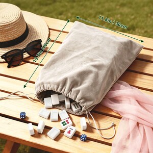 May include: A beige drawstring bag, 11.8 inches wide and 15.7 inches tall, spills out white game tiles with red and green markings. A straw hat and black sunglasses rest on a wooden table. Two blue dice and a pink scarf are also visible.