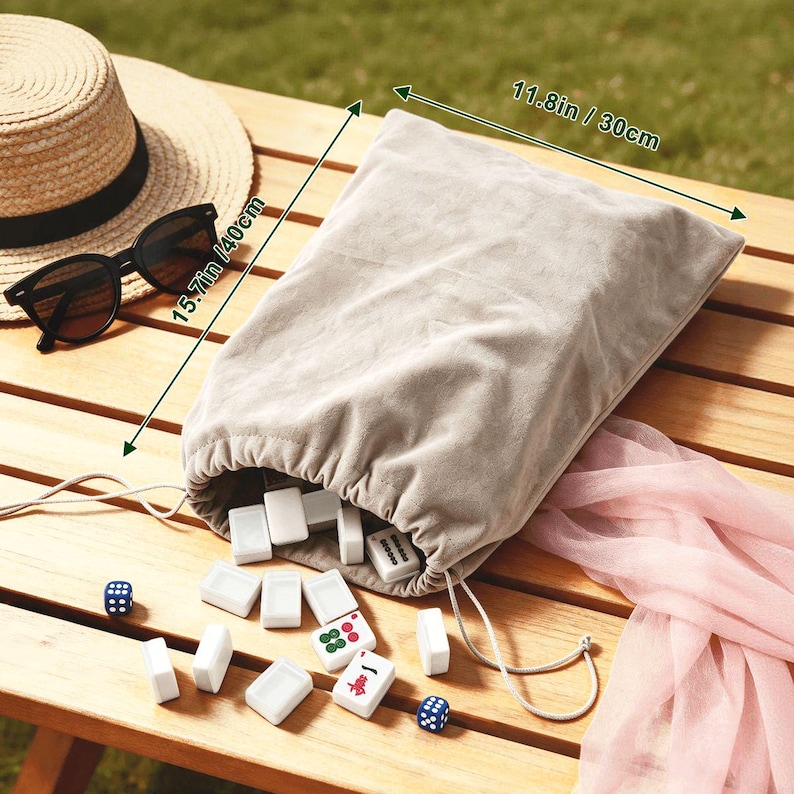 May include: A beige drawstring bag, 11.8 inches wide and 15.7 inches tall, spills out white game tiles with red and green markings. A straw hat and black sunglasses rest on a wooden table. Two blue dice and a pink scarf are also visible.