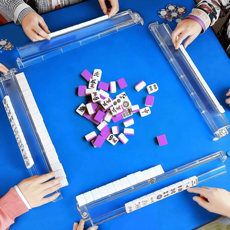 May include: A group of people playing Mahjong on a blue table. The game features white and purple tiles, clear plastic tile racks, and game chips. The tiles are scattered in the center of the table, with players arranging them in front of them.