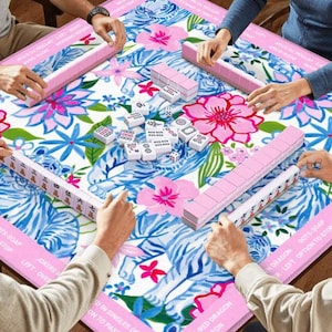 May include: A colorful Mahjong game in progress. The game board features a floral and tiger stripe design in pink, blue, and white. Pink tiles and racks are visible, with players' hands around the table. The game is set up for play.