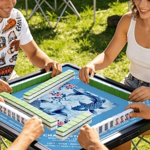 Peut inclure: Une partie de Mahjong en cours, jouée par plusieurs personnes. Le plateau de jeu est bleu avec un motif de carpes koï. Des tuiles vertes et blanches sont disposées autour du plateau. Le texte sur le plateau indique "CHA RIGHT ST ER-LE OPTION TO STOP LEFT - OVER - RIGHT OPTION TO PASS 1-3 TILES."