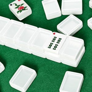 May include: White Mahjong tiles with green and red symbols are scattered on a green surface. The tiles are rectangular and have various symbols and numbers. Some tiles are stacked, while others are separate. The game pieces are ready to be played.