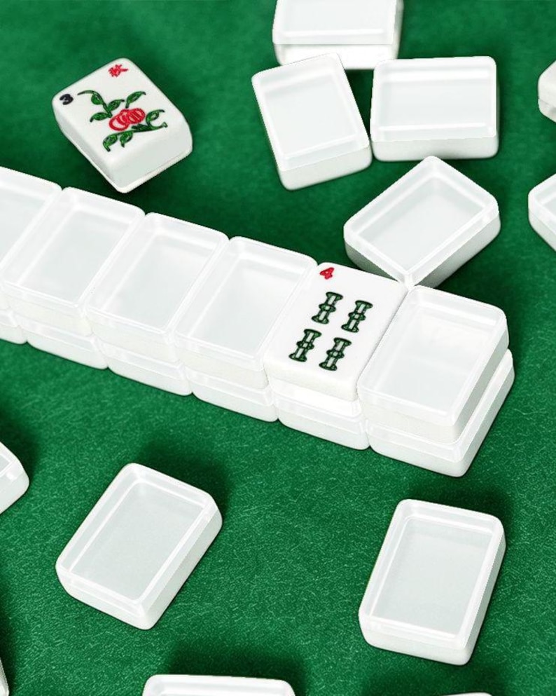May include: White Mahjong tiles with green and red symbols are scattered on a green surface. The tiles are rectangular and have various symbols and numbers. Some tiles are stacked, while others are separate. The game pieces are ready to be played.