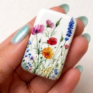 May include: A rectangular, white ceramic tile featuring a watercolor floral design. The artwork showcases vibrant blooms in shades of pink, red, yellow, and blue, with green stems and leaves. The tile is held in a hand.