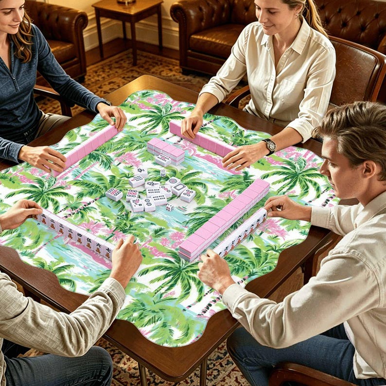 Op de afbeelding: Een groep mensen speelt mahjong aan een houten tafel. De tafel is bedekt met een tropisch thema doek met palmbomen en roze bloemen. De mahjong-tegels zijn wit met zwarte markeringen en de rekken zijn roze.