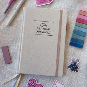 May include: A beige Reading Journal with the title printed in black, a pink elastic band, and several stickers. Stickers include a pink heart with text, a pink card with cherries, and a pink sticker with text. Two pens and a color swatch are also visible.