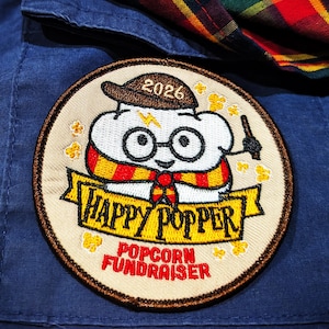 May include: A circular embroidered patch featuring a white popcorn character wearing glasses, a scarf, and a hat labeled 2026. The patch includes the text HAPPY POPPER and POPCORN FUNDRAISER in red and black, set against a blue fabric with a plaid pattern.