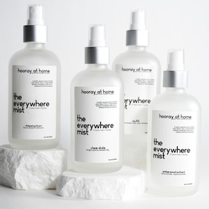 Pode incluir: Quatro garrafas de spray de vidro transparente com tampas prateadas e etiquetas brancas. As etiquetas dizem "hooray at home" e "the everywhere mist" com diferentes aromas: "whipering bloom", "clean slate", "hot oil" e "amberwood orchard".