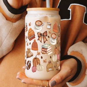 May include: A reusable coffee cup with a brown, white, and orange design featuring fall-themed illustrations like pumpkins, leaves, and coffee cups. The cup is made of stainless steel and has a lid.