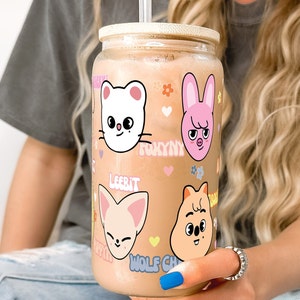 May include: A clear glass tumbler with a bamboo lid and a straw. The tumbler is filled with a light brown liquid and has colorful cartoon animal stickers on the outside. The stickers include a white cat, a pink bunny, a yellow fox, and a brown bear. The text on the stickers reads 'LeeBit', 'Tokyny', 'Wolf Ch', and 'Happin'.