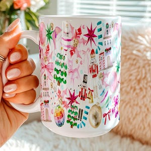 May include: A white ceramic mug featuring a colorful watercolor pattern of Christmas-themed illustrations, including nutcrackers, decorated pine trees, pink bows, stars, ornaments, and small buildings, all arranged in a festive holiday design.