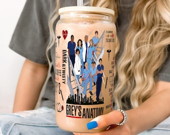 May include: A reusable tumbler with a brown lid and a straw. The tumbler features a graphic of the main characters from the TV show Grey's Anatomy, along with the show's title and the phrase "Dark & Twisty".