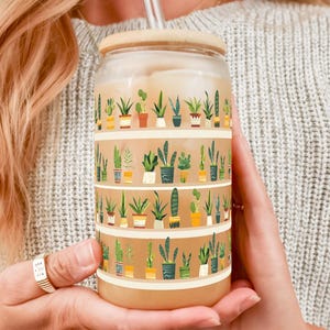 May include: A clear glass reusable cup with a brown wooden lid and a straw. The cup has a pattern of potted plants in various colors and styles.