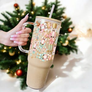 May include: A tall, clear glass tumbler with a brown lid and a straw. The tumbler is decorated with a pattern of pink and blue nutcracker figures on a white background. The tumbler is filled with a light brown liquid.