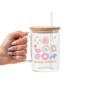 May include: A clear glass mug with a bamboo lid and a straw. The mug is decorated with colorful summer-themed illustrations, including a flamingo, donut, and rainbow. The text "GO WITH THE FLOAT" is printed on the mug.