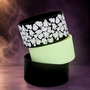 May include: A stack of three cylindrical silicone sleeves. The top sleeve is black with a repeating white ghost pattern. The middle sleeve is a solid pale lime green. The bottom sleeve is a solid black. Each sleeve has a smooth, matte finish.