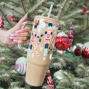 May include: A clear glass tumbler with a wooden lid and a straw. The tumbler has a festive design featuring nutcracker figures, candy canes, and snowflakes. The tumbler is filled with a light brown beverage.